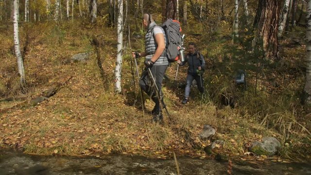 People walk near a mountain stream. Family travels. People environment by mountains, rivers, streams. Parents and kids walk using trekking poles. Man and woman have hiking backpacks, flasks, mugs. Dad
