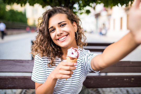 Portrait of young woman eating ice cream and taking selfie photo on camera in summer street.