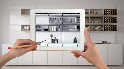 Hands holding and drawing on tablet showing minimalistic white and wooden kitchen CAD sketch. Real finished interior in the background, architecture design presentation, top view