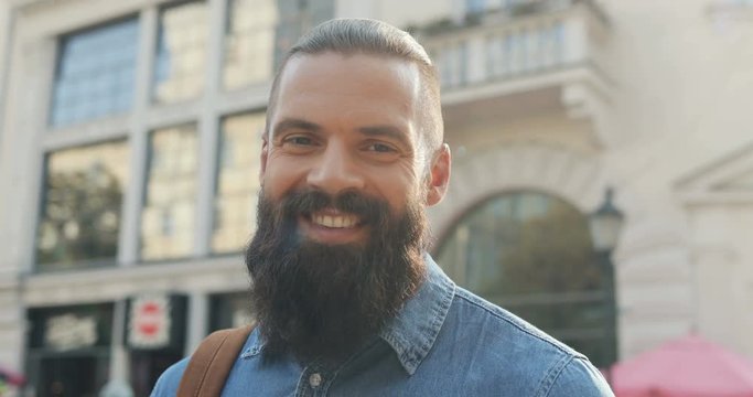 Portrait of the handsome Caucasian man with a beard looking at the side and then smiling to the camera at the street of a town. Close up. Outdoors.