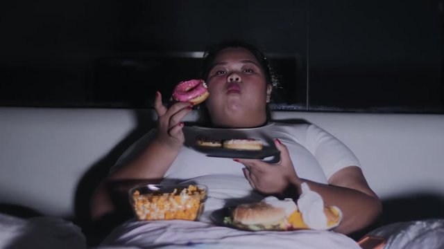 Overweight woman eats donuts and others junk foods on the bed at night before sleep at home. Shot in 4k resolution