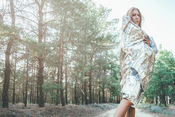 Fototapeta na wymiar Beautiful girl is wrapped in a foil emergence blanket. Wet model stands in the forest in the morning. Concept of the tourist who studied the problems of pollution of nature who was wet from the water