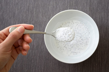 Hand holding the spoon of the plate of chia seeds with coconut milk, grey tablecloth background, top view