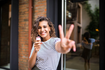 Carefree and happy, sunny spring mood. Cute young curly girl eating ice cream and making peace gesture on camera on the city street