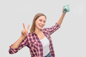 Obraz premium Young woman holding blue camera in hand and stretch arm up. She poses and smiles. Woman looks straight. She shows piece symbol with fingers. Isolated on grey background.