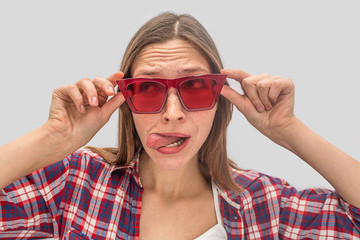 Funny young woman stands close and looks to left through red sunglasses. She holds it with both hands and shows tongue. Woman is playing role. Isolated on grey background.