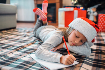 Calm and peaceful girl lying on floor and writing letter. She keeps her legs crossed and holding head on hand. Child is in room alone. She wears Christmas hat.