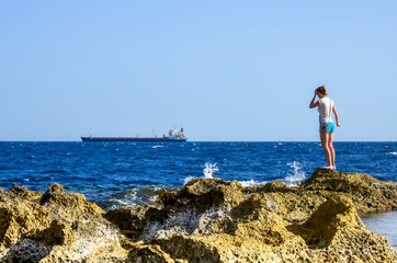 Woman on a rocky beach with an oil tanker in the distant sea horizon. Photo taken in the Mediterranean Sea, Malta