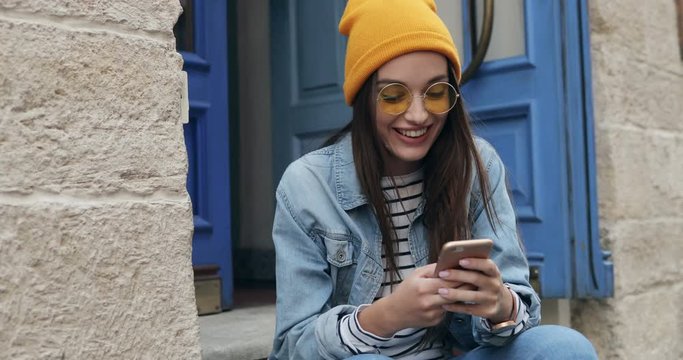 Pretty young hipster girl in sunglasses and hat sitting on the steps of the door and chatting on the smartphone and smiling. Portrait shot. Outdoors.