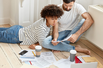 Family couple maintains home accounting, discuss plans and future payments, surrounded with bills on floor, dressed in domestic clothes, drinks tasty hot coffee, analyze income chart together
