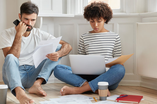 Photo of two mixed race coworkers read documentation, concentrated on solving problem with planning, check email box on laptop computer, drink coffee, look through delivered bills, sit on floor