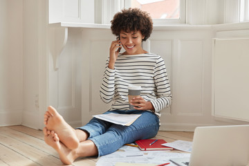 Glad black woman with Afro haircut, talks via cell phone while reviews documents, uses laptop computer during coffee time, wears striped sweater and jeans, has bare feet, sits on floor in empty room