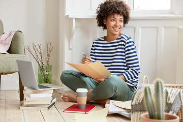 Positive black woman sits crossed legs, dressed in striped sailor sweater, holds some papers busy with preparing report, learns scientific literature, uses laptop, drinks hot beverage, works from home