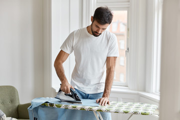 Hard working bearded man dressed in white t shirt, irons shirt on ironing board, takes care of clothes, does household duties, poses against window background. Housework and masculinity concept - obrazy, fototapety, plakaty