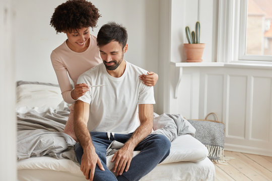 Happy young African American woman shows positive pregnancy test to her husbnad, rejoice good news of becoming parents soon, pose on unmade bed in spacious cozy bedroom. Planning family concept