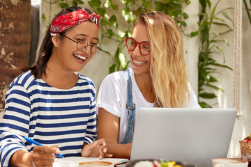 Horizontal view of cheerful mixed race women write article about distant job, learns language by internet, wear round spectacles, drink coffee, have joyful expressions, write notes in notebook
