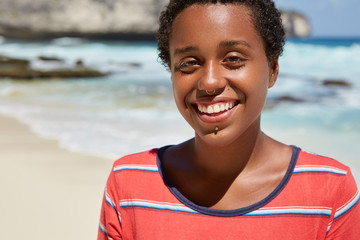 Headshot of pleasant looking cheerful black young woman with boyish appearance, smiles broadly, shows white perfect teeth, dressed in striped casual t shirt, poses against blurred blue ocean view