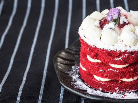 Close Up Of Homemade Red Velvet Cake (chocolate Cake With Red Food Coloring), Fresh Cream And Berries On Top, On A Black Plate And On The Table Covered With A Black And White Stripes Tablecloth.