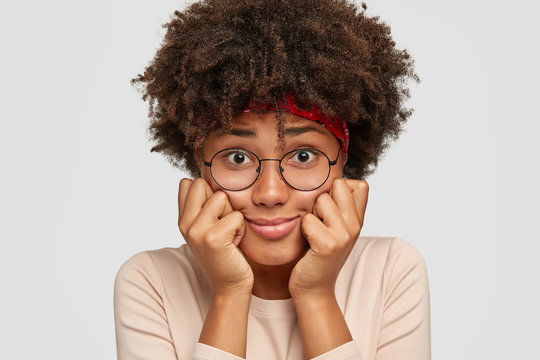 Close up shot of embarrassed black lady has surprised dejected facial expression, keeps hands on cheeks, has Afro haircut, models against white background, feels lonely. Emotions and lifestyle concept