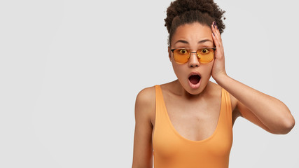 No way, its shocking! Surprised black woman with curly hair, wears sunglasses, keeps jaw dropped, dressed casually, poses against white background with free space for your advertisement or text