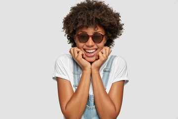 Headshot of smiling pleased African lady, has bushy hair, happy expression, holds chin with both hands, wears trendy shades, white t shirt and overalls, ready to have picnic with friends poses indoor