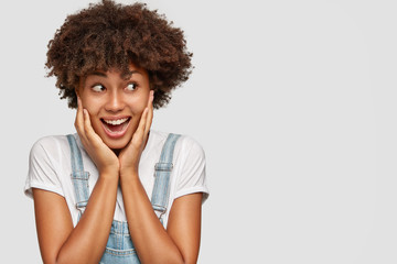 Positive young girl with dark skin, keeps hands on cheeks, looks happily aside, notices nice place...