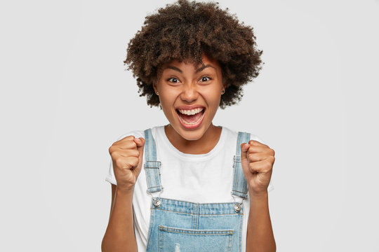 Photo of annoyed outraged black young lady clenches fists with anger, has Afro hairstyle, wears denim dungarees, expresses negative emotions, models over white background, shouts desperately