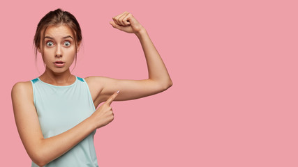 Horizontal studio shot of surprised European girl indicates at her muscle, stares at camera with eyes popped out, cant believe in such quick results, has outdoor trainings on biceps, likes sport