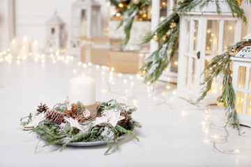 Christmas candles and snowy fir branches over white wooden background with lights.  New Year's decoration with a fir tree in white tones.