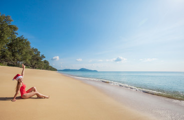 Fototapeta na wymiar Happy young woman with long hair in red swimsuit and santa claus hat sitting on the wide tropical beach with no people near the sea with beautiful blue water and sky on Phuket island,Thailand