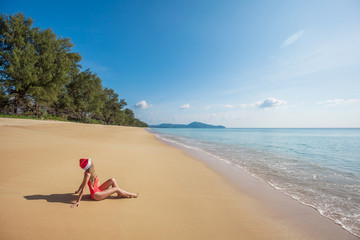 Fototapeta na wymiar Young woman with long hair in red swimsuit and santa claus hat sitting on the wide tropical beach without people near the sea with beautiful blue water and sky on Phuket island,Thailand
