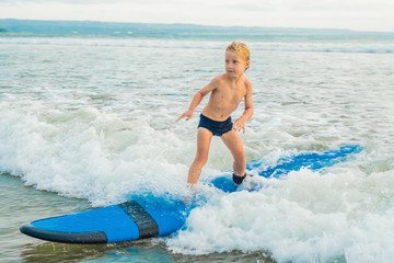 Little boy surfing on tropical beach. Child on surf board on ocean wave. Active water sports for kids. Kid swimming with surf. Surfing lesson for kids