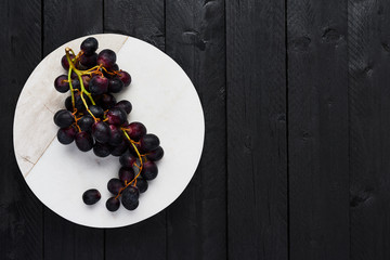 Top view of fresh ripe black grapes on white marble serving plate on black wooden table with copy space.