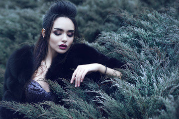 Young beautiful lady with perfect provocative make up and combed hairstyle wearing luxurious sequin dress and black sable fur coat sitting in coniferous bush and touching it with her fingers