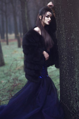 Young beautiful lady in luxurious sequin evening dress and sable fur coat standing in the mysterious misty woods leaning on the moss covered tree trunk with enigmatic look