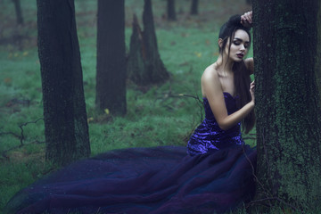 Young beautiful lady in luxurious sequin evening dress sitting in the mysterious misty woods leaning on the moss covered tree trunk with sorrowful look