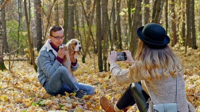 Girl trying to take a photo of a guy with a dog, that twisting out his hands, on mobile phone in autumn forest