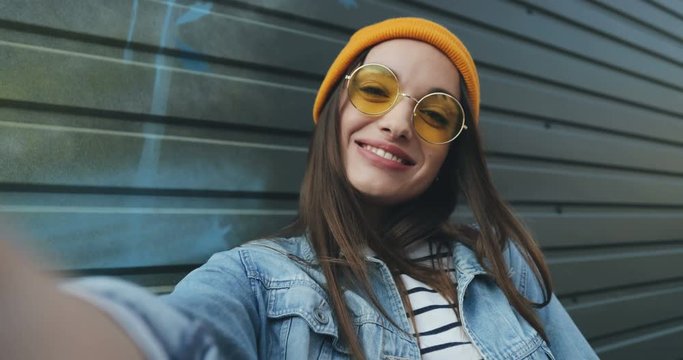 Close up of the stylish Caucasian cheerful girl in sunglasses and hat taking cool selfie pictures and posing with rock sign. Outdoor. POV. Portrait.