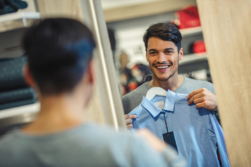Young man choosing shirt and looking to mirror in mall or clothing store