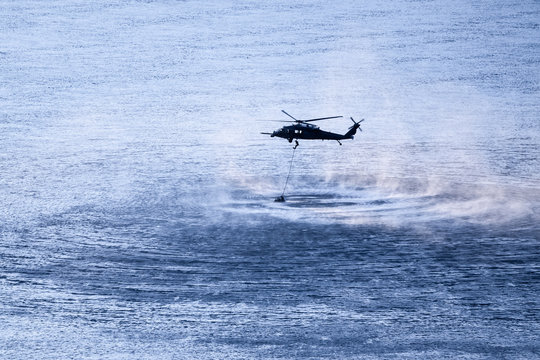 Military Helicopter Performing Training Rescue Operation Over The Columbia River At Sunset. Water Splashes, Vapor And Sprays Rise From The River. Rope Hangs From The Helicopter With Diver Sliding Down