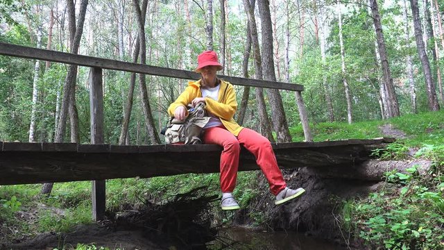 Mature Female traveler sits on a bridge over summer forest stream and takes pictures and talks through a video call on phone