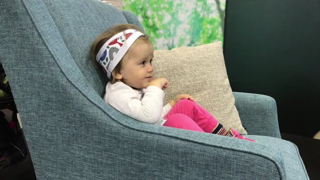 Little baby girl sitting in armchair looking away, smiling and pointing a finger at something. Toddler raptly watching TV and vocalising with emotions. Child interest to cartoon or moving pictures