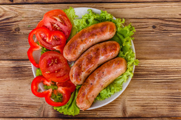 Ceramic plate with grilled sausages, sliced tomatoes and lettuce leaves on wooden table. Top view