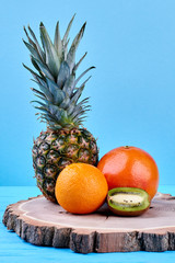 Composition from exotic fruits on tree stump. Ripe ananas, grapefruit, orange and kiwi fruit on blue background. Fresh dietary food for sportmans.