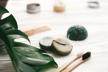 Eco friendly natural bamboo toothbrushes, shampoo bar, toothpaste in glass, wooden brush and konjaku sponge on white wood with green monstera leaves. Zero waste concept, plastic free