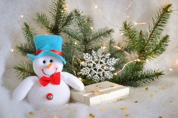 Funny felt snowman with a gift on the background of fir branches with lights of garland on the snow on a festive day.