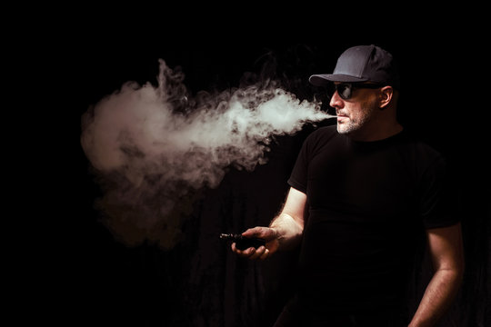 Vaper. A man produces smoke. E-Sigs. A man smokes electronic cigarettes. Vaper on a black background. Vaping man holding a mod. A cloud of vapor. Vaping. e-cigarette