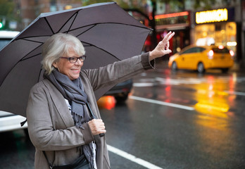 Mature white haired woman hailing taxi cab on New York city street under umbrella on rainy day