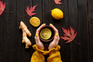 Mug of tea in female hands. Lemon, ginger and autumn leaves. Cold treatment. Wooden background. Top...