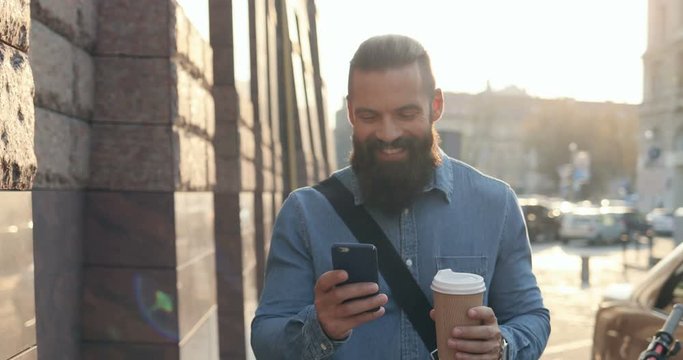 Attractive Caucasian young hipster man with a beard chatting on the smartphone cheerfully and smiling while stepping the town with a coffee in a hand. Outdoors.